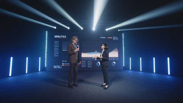 An adult businessman on the stage in casual clothes telling the female presenter and the audience about the graphs and company finance on the LED screen in studio, during a business training lecture