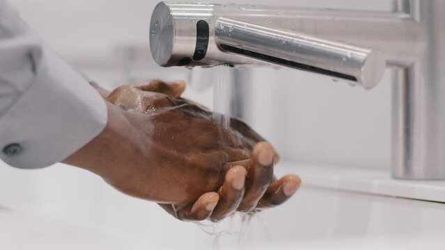 Close-up young unrecognizable african american man businessman washes hands in modern bathroom slow motion rinsing dirty hands under warm water over sink concept daily hygiene protection from viruses