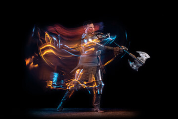 Obraz na płótnie Canvas Dynamic portrait of brutal serious man, medieval knight with swinging ax fighting isolated over dark background in neon mixed light. Art, history, creativity