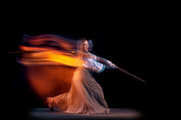 Portrait of beautiful woman in image of medieval warrior or knight with dirty wounded face with big sword isolated over dark background in mixed neon light.