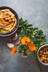 vista superior de un bol con hummus y un bol con garbanzos, acompañados de ajos, perejil y flores...