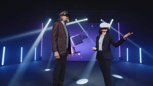 Two presenters in suits on stage trying new augmented reality glasses at a Metaverse conference in a studio with spotlights in front of LED screen, with 3D objects