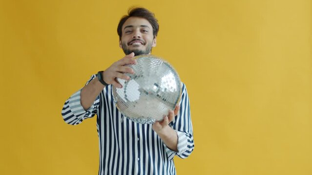 Slow motion portrait of happy Indian guy dancing holding disco ball moving on yellow color background. Handsome brunet wearing trendy clothing enjoying party alone.