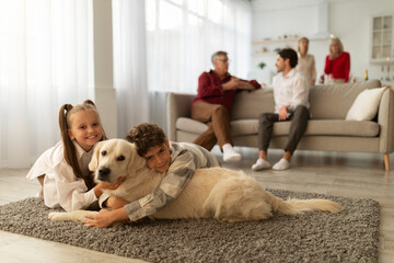 Happy kids hugging their golden retriever, lying on floor together, their relatives communicating on couch at home