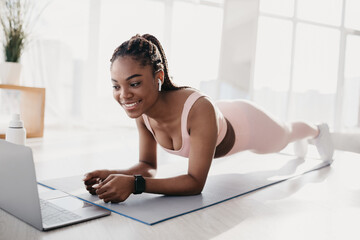 Sporty millennial black woman standing in elbow plank, following online video tutorial on laptop...