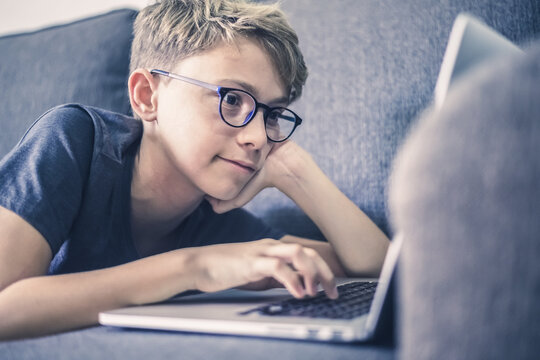 Young student doing homework lying on a sofa using laptop. Caucasian boy watching video online alone at home. Teen enjoying free time with computer. New technology education concept Soft focus on eyes