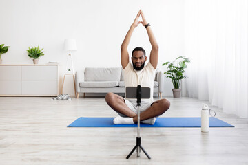 Calm millennial black bearded man in white sportswear sits on mat in lotus position, practices yoga