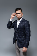 bearded businessman in blazer adjusting glasses and looking at camera isolated on grey.