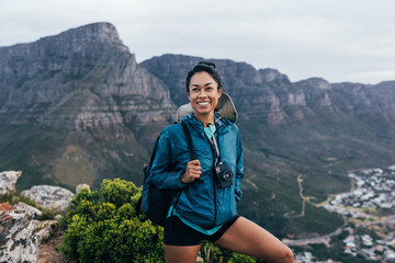 Portrait of a young woman hiker wearing an analog camera and backpack standing on a top of a...