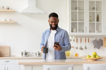 Cheerful young black male with cup of coffee in hand in minimalist kitchen interior reads news on smartphone
