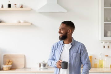 Happy young bearded african american guy with cup of coffee in hand in minimalist kitchen interior looks at empty space