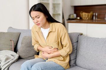 Concerned asian woman suffering from stomach-ache sitting on sofa. Female holding tummy, undergoing...