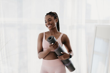 Portrait of young pretty black woman in sportswear holding fitness mat, posing and smiling at home...