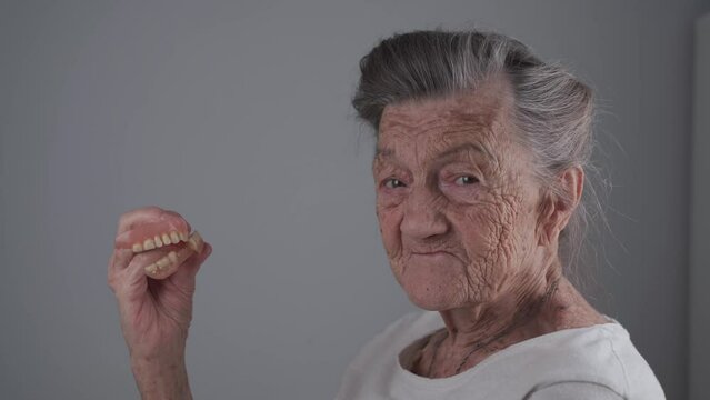 Elderly woman took off removable dentures, shows to camera and smiles. Oral care concepts, false teeth and old age. Full removable denture in hands of old female with gray hair studio video filming.