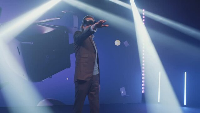 Excited presenter on stage gesturing and demonstrating new augmented reality glasses to the audience, at a Metaverse event in a large room with spotlights in front of an LED screen with 3D objects