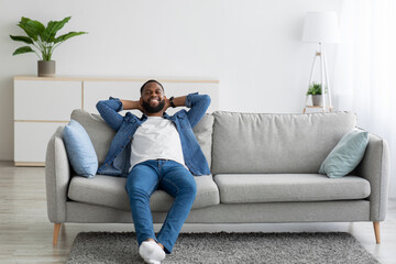 Satisfied relaxed handsome young african american bearded guy resting on sofa in living room...
