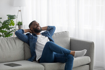 Fototapeta premium Happy satisfied relaxed young black bearded male resting in living room interior with tablet on sofa