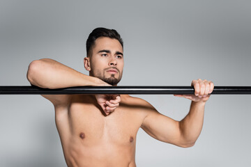 muscular sportsman looking away and leaning on horizontal bar isolated on grey.