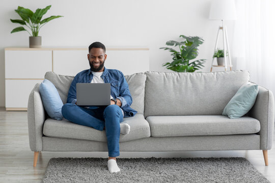Smiling millennial black bearded guy working on laptop on sofa in living room interior