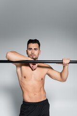 muscular sportsman looking at camera and leaning on horizontal bar on grey.