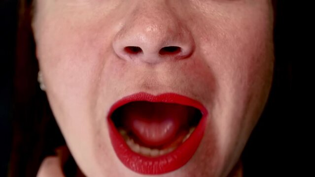 Close-up of the face of a caucasian woman with lips painted with red lipstick sings song, opening her mouth wide on a black background.