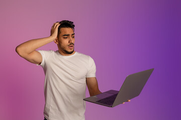 Shocked young Arab man with laptop computer touching his head in panic, missing work project deadline in neon light