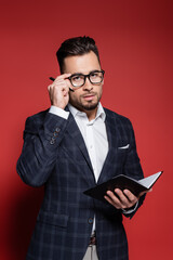 bearded businessman in checkered suit adjusting glasses while holding pen and notebook on red.