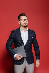 Fototapeta premium bearded businessman in blazer and glasses holding laptop on red.