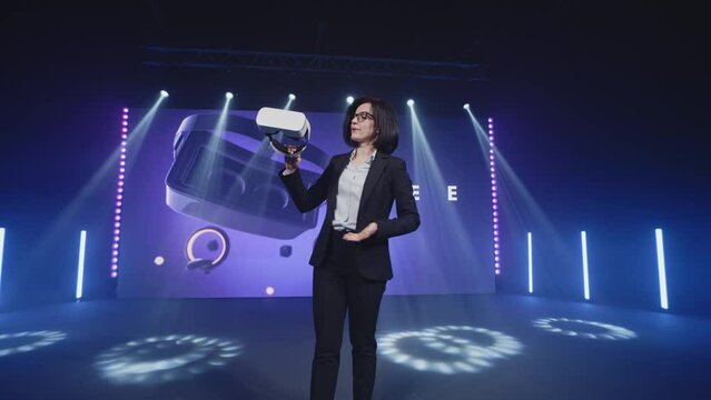 A female presenter standing on stage in modern studio with LED screen and holding a new model of augmented reality glasses, during a Metaverse conference and speaking about a new haptic technology