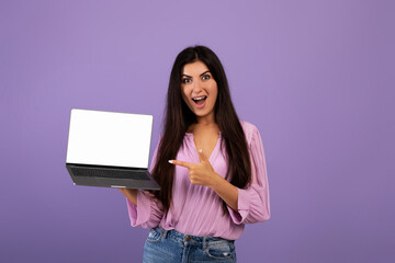 Excited armenian young woman pointing at brand new notebook with blank display, posing on violet background, mockup