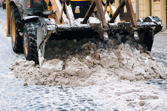 An Excavator Bucket Shovels Snow From Paving Stones On A City Street In Lviv, Ukraine. The Concept Of Dealing With The Consequences Of Snowfall.