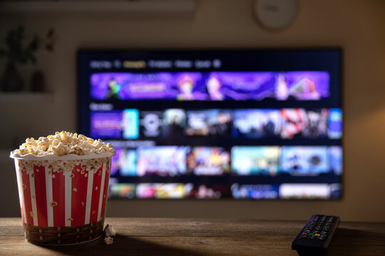 Close up of bowl of popcorn and remote control with tv works on  background. Evening cozy watching a movie or TV series at home.