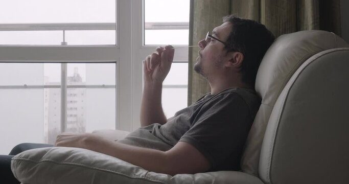 Man uses nasal swab to test for Covid-19 at home, sitting in white leather armchair near window. Coronavirus, self rapid antigen test, side view, profile.