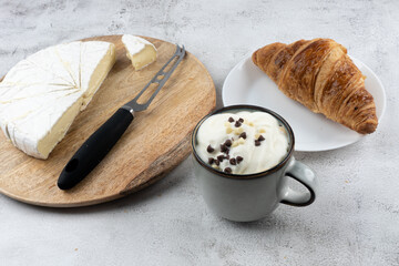 A cup of coffee, Butter bun, cheese knife and sliced round white cheese - on a wooden board plate. HOME breakfast