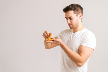 Portrait of confused young man with disgust picking bud of spoiled burger on white isolated background. Studio shot of handsome bearded male holding in hands unhealthy cheeseburger.