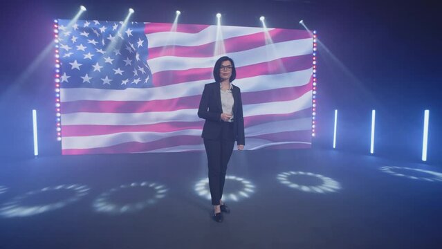 Female presenter in suit on stage talking about the latest political and public news of the United States on TV broadcast in the modern studio, with spotlights near the American flag on the LED screen