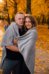 Fototapeta premium Young couple is walking in the sunny autumn park. Happy boy and girl are rolling themselves in the rug near yellow tree. Fallen yellow leaves, brith warm autumn weather. Autumn love story in park 