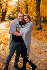 Obraz premium Young couple is walking in the sunny autumn park. Happy boy and girl are rolling themselves in the rug near yellow tree. Fallen yellow leaves, brith warm autumn weather. Autumn love story in park 