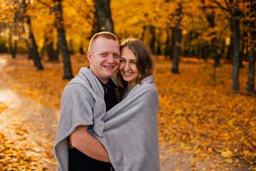 Fototapeta premium Young couple is walking in the sunny autumn park. Happy boy and girl are rolling themselves in the rug near yellow tree. Fallen yellow leaves, brith warm autumn weather. Autumn love story in park 