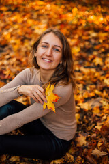 Beautiful girl is sitting on yellow leaves in the park. Attractive woman wearing beige sweater and black pants is walking in autumn park. Bright, warm sunny autumn day with yellow background.
