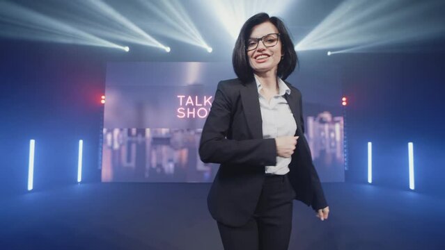 Happy presenter in a suit and glasses running onto the stage and announcing the start of the famous comedy, late-night show in an illuminated room with LED screen and 3D inscription