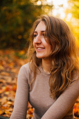 Fototapeta premium Beautiful girl is sitting on yellow leaves in the park. Attractive woman wearing beige sweater and black pants is walking in autumn park. Bright, warm sunny autumn day with yellow background. 