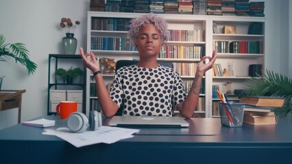 Young peaceful African American woman office worker meditates at workplace doing breathing practices closing eyes doing yoga pose with hands sits on chair at table. Relaxation, stress relief
