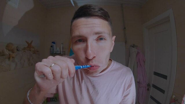 A young man in a pink T-shirt brushes his teeth in front of a mirror.