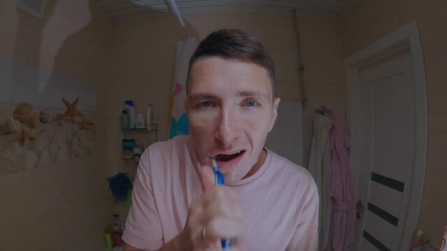 A young man in a pink T-shirt brushes his teeth in front of a mirror.