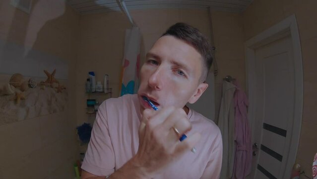 A young man in a pink T-shirt brushes his teeth in front of a mirror.