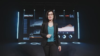 Successful businesswoman in a casual suit and glasses, announcing to the audience the start of a business training in modern room near LED screen with business analytics charts and growth percentages
