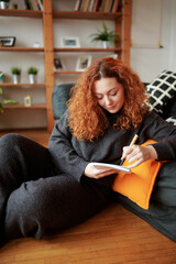 A happy cute ginger girl with curly hair sits on the floor at home and writes notes in a notebook.