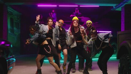 Group of stylish young people dancing , moving near military tank inside garage building Colorful stylish clothes , hippie style fashion . Modern dances . Purple led light , slow motion