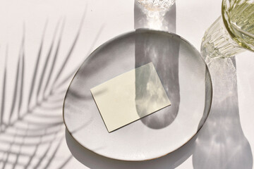 Eco minimal tableware scene.  Empty blank on ceramic plate, cocktail glass on neutral background. Sunlight, palm shadow.  Mockup invitation card for wedding, business or menu. Wabi sabi style